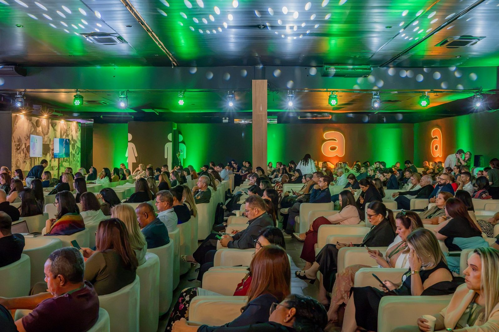 Público sentado em poltronas claras acompanhando a palestra