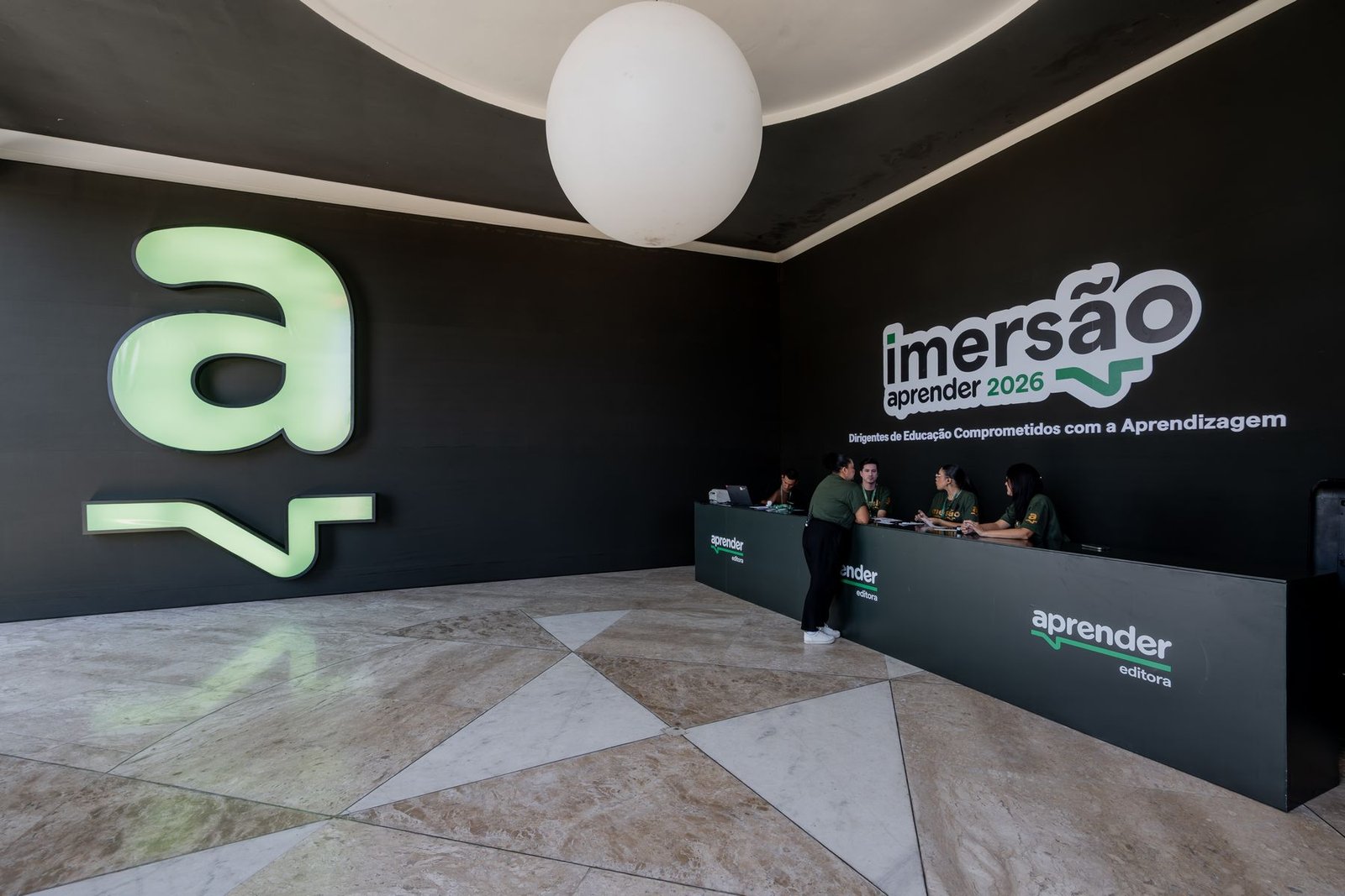 Recepção da Imersão Aprender: letra a iluminada em verde na parede e balcão de credenciamento