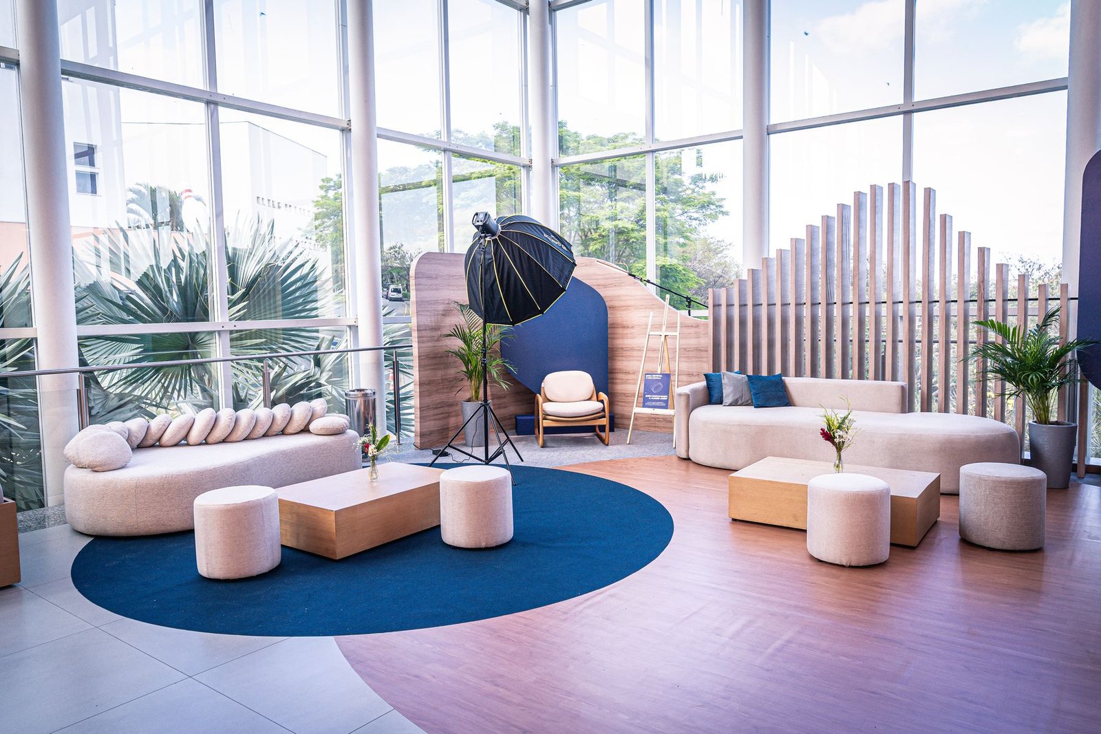 Lounge do Encontro de Mantenedores: sofás curvos claros, tapete azul circular, painel ripado de madeira e pé-direito envidraçado