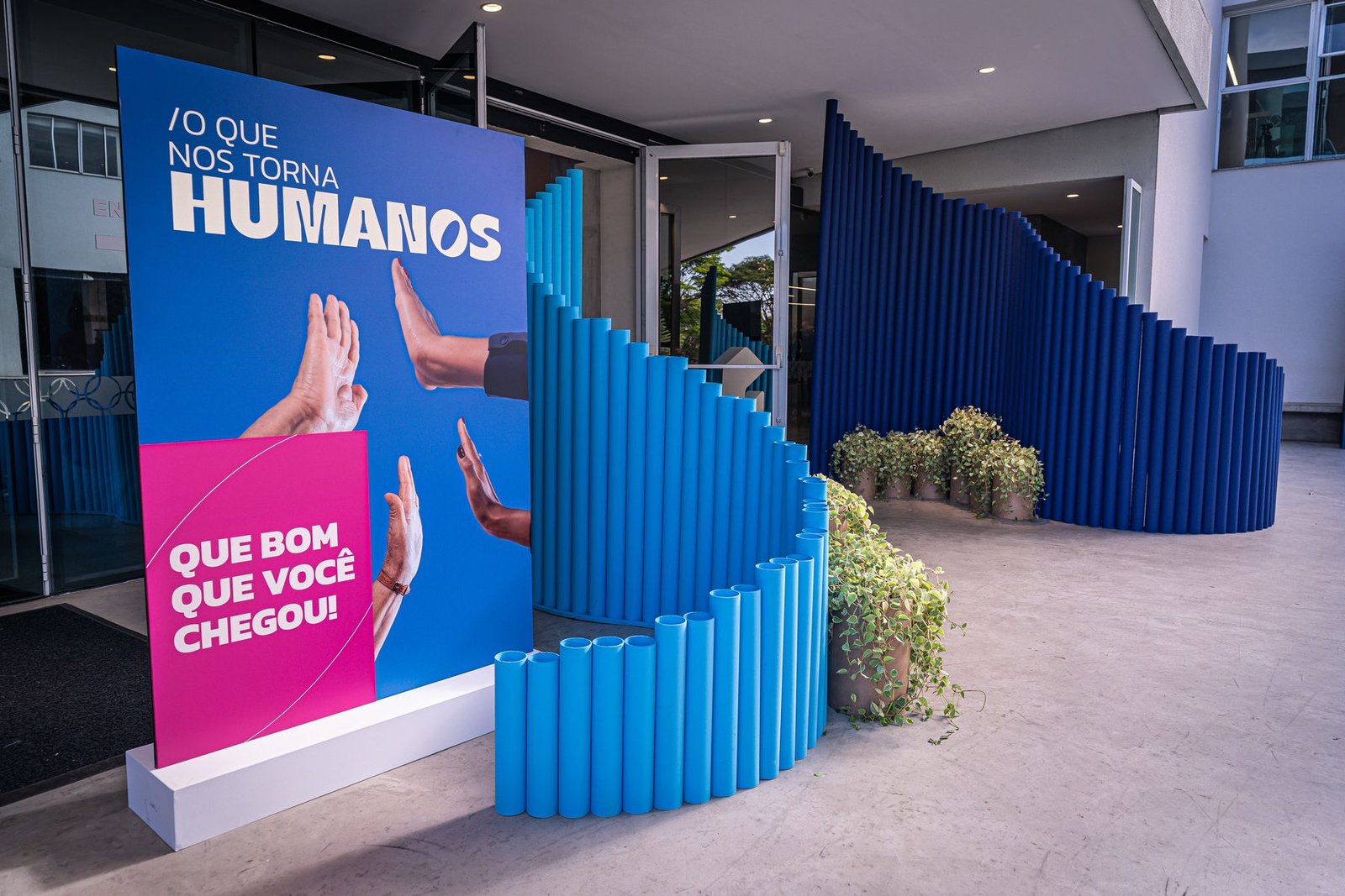 Totem de boas-vindas em azul e rosa junto à parede de tubos