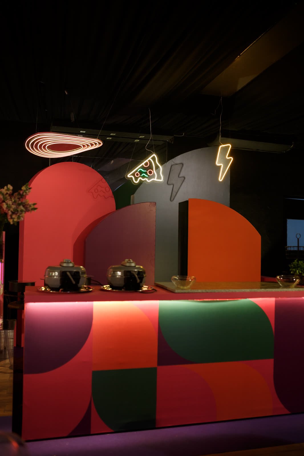 Balcão cenográfico em laranja, rosa e verde com neon de raio e arcos ao fundo