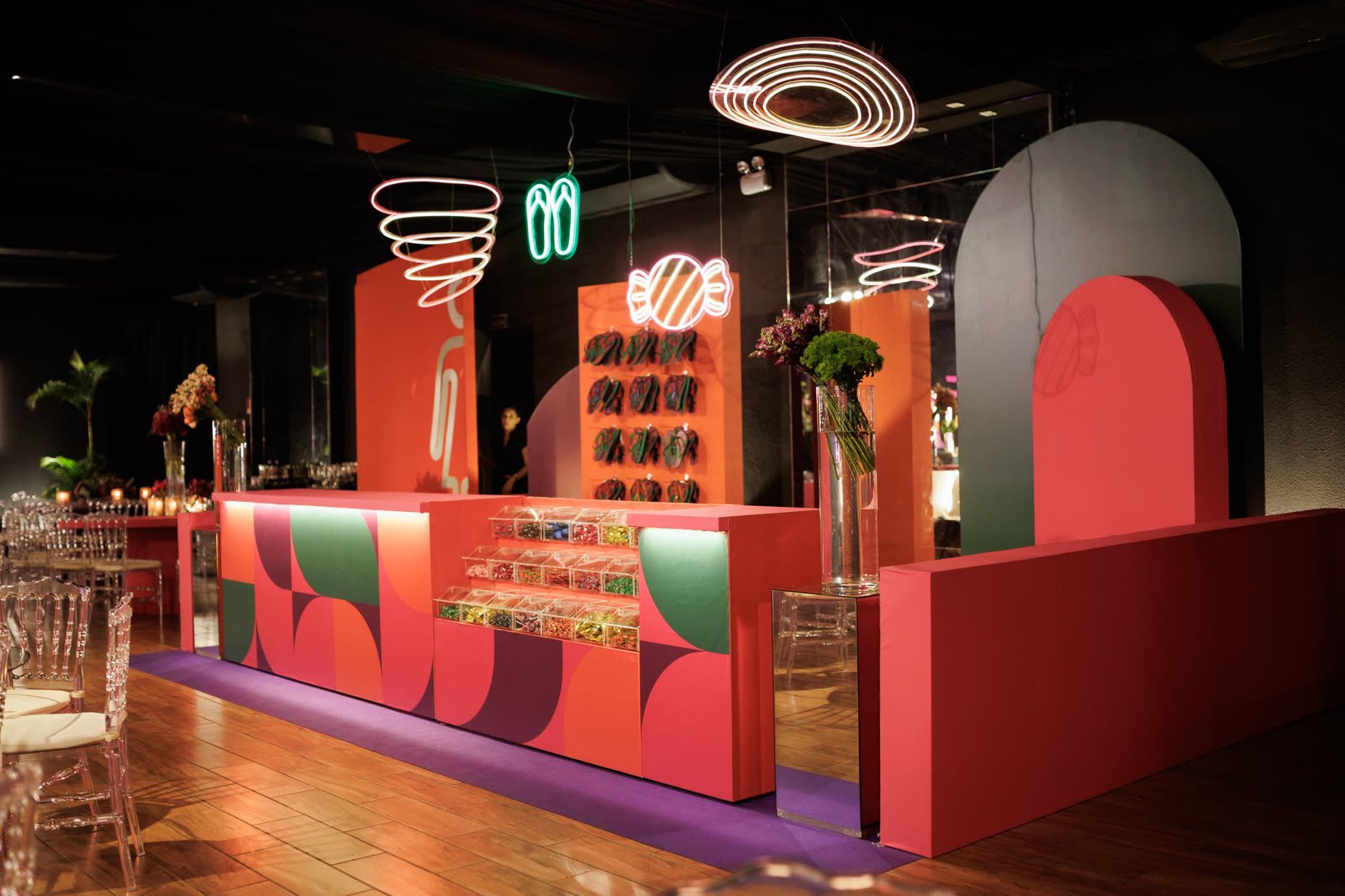 Bar em blocos de laranja, verde e roxo, com neons de doces e chinelos e arcos cenográficos