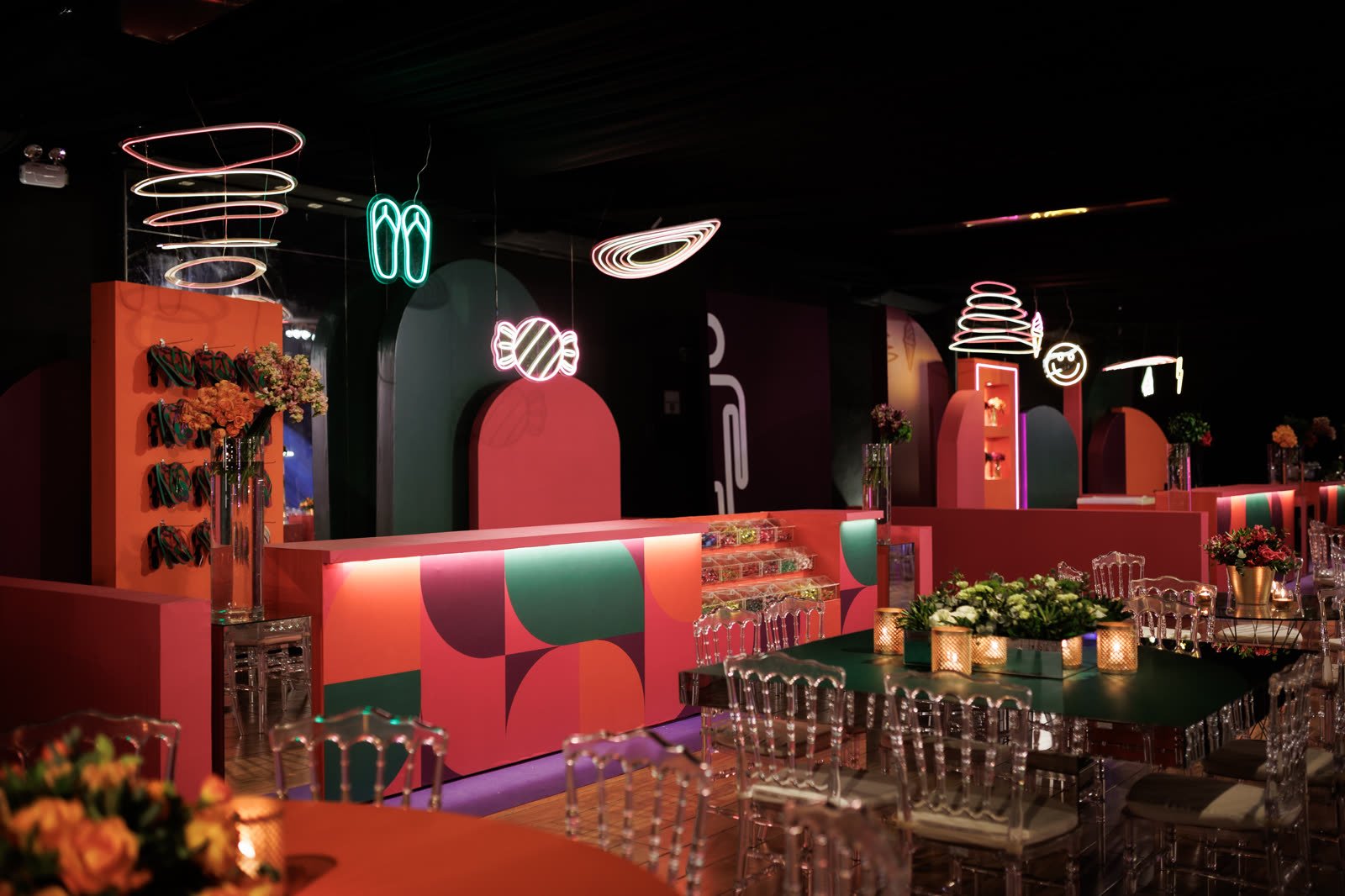 Lounge e bar com neons, arcos laranja e verde e mesas com arranjos de flores
