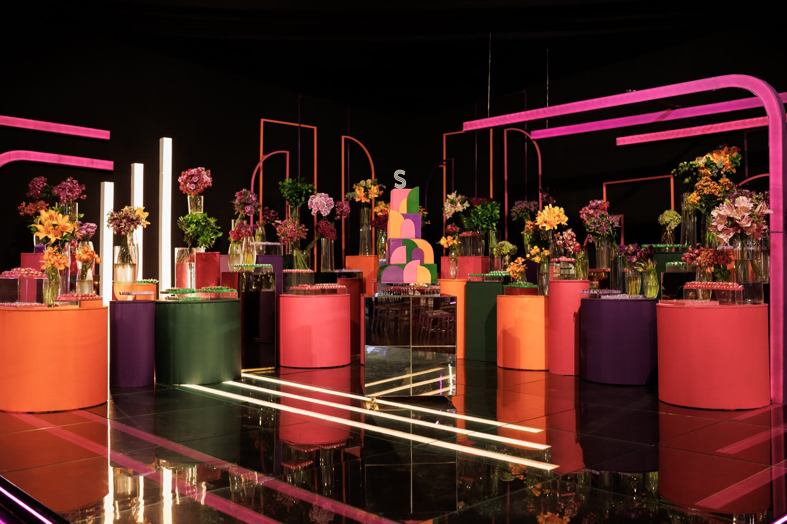 Mesa do bolo sobre cilindros coloridos, flores e estruturas em neon rosa, com piso espelhado