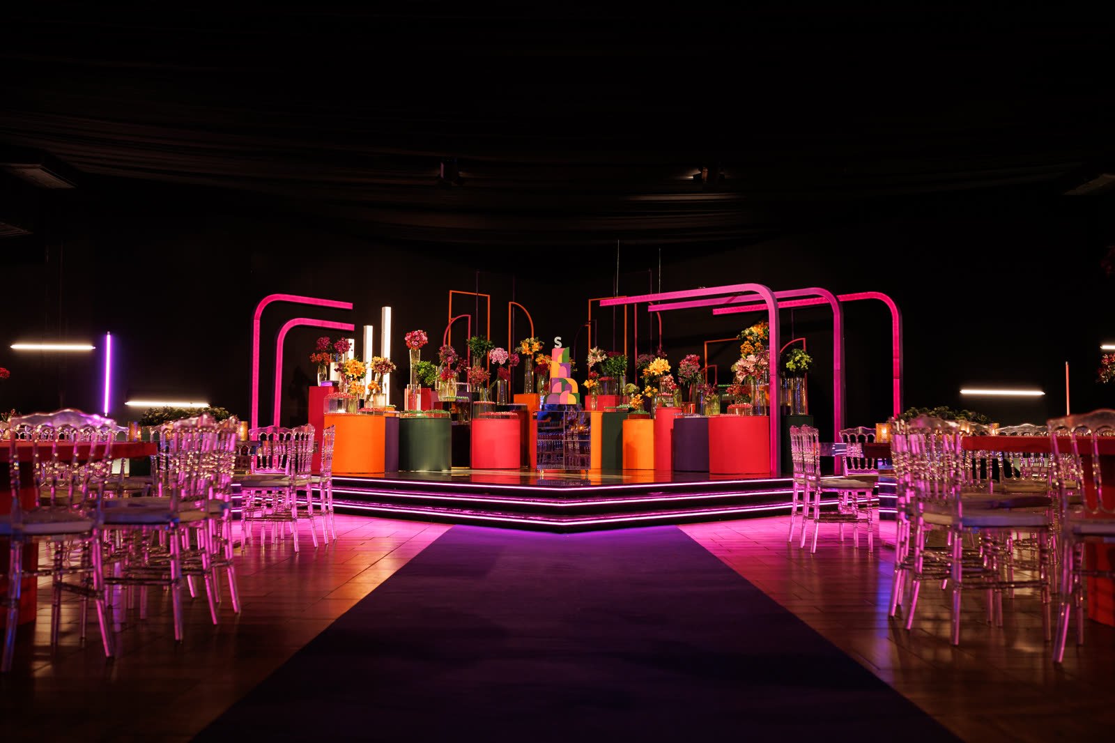 Salão de festa de 15 anos com pista em neon rosa, mesas com cadeiras de cristal e mesa do bolo cercada de flores