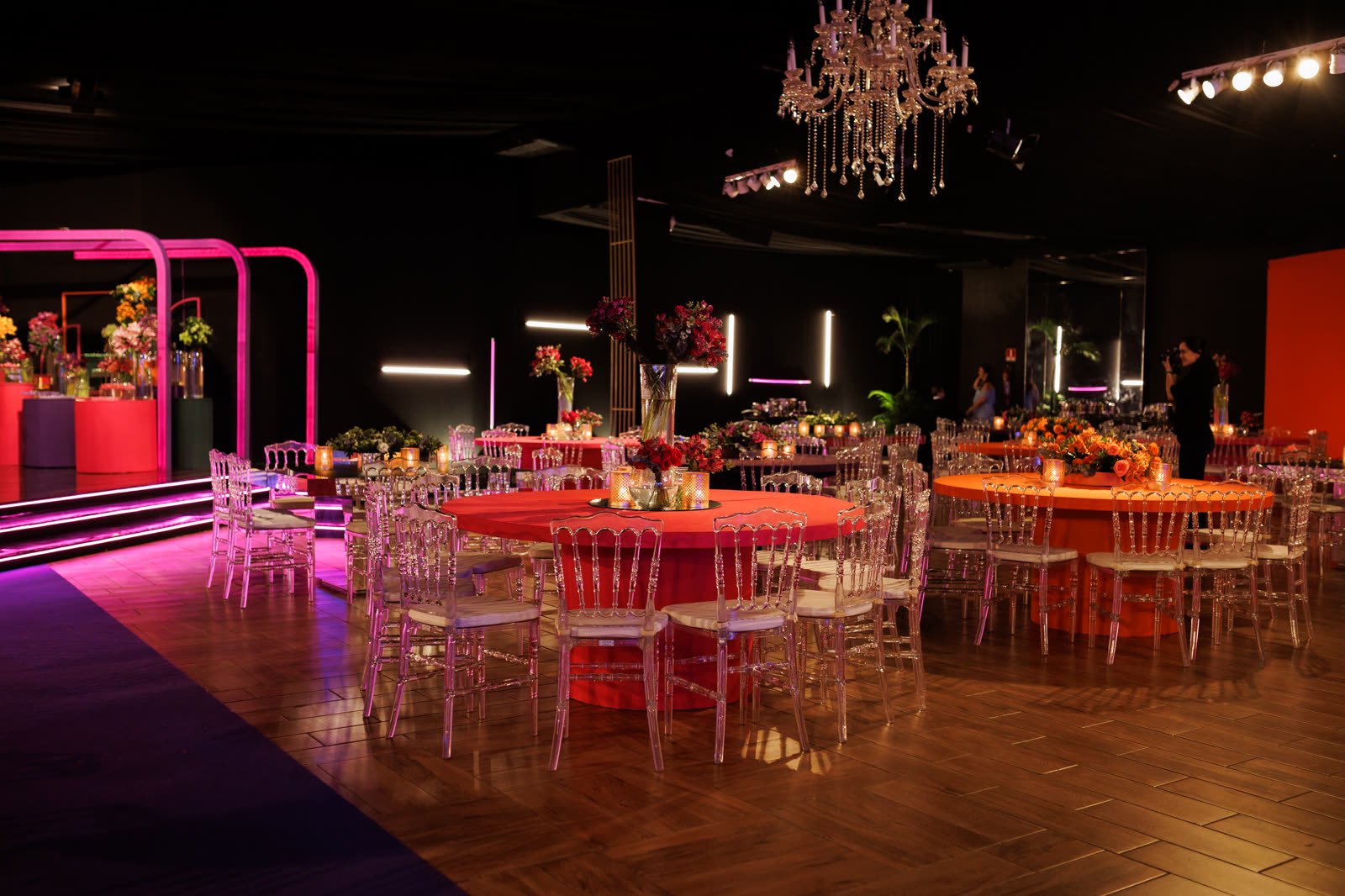 Salão com lustre de cristal, mesas redondas laranja e vermelhas e a pista de neon