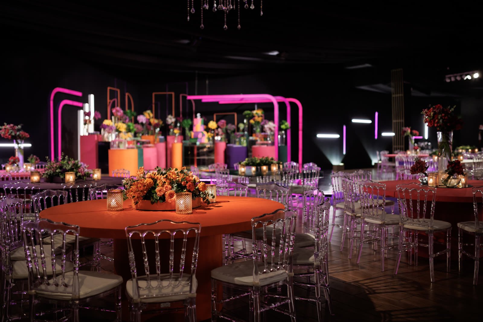 Mesa redonda laranja com arranjo de rosas, cadeiras de cristal e a pista de neon ao fundo